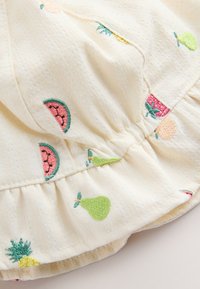 Krämfärgat tyg med broderade färgglada fruktmotiv inklusive vattenmelon, päron, ananas och jordgubbe på en samlad volangkant.