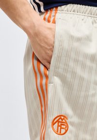 Mână în buzunarul pantalonilor sport bej cu dungi portocalii și logo brodat, purtați cu o cămașă albastru închis cu margine albă.