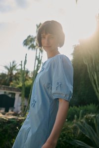 Jeune femme aux cheveux courts portant une robe bleu clair, debout dehors près de grands cactus avec la lumière du soleil qui brille derrière elle.