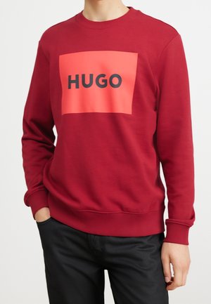 Osoba nosící červenou mikinu s velkým oranžovým čtvercem a černým nápisem "HUGO" přes hruď, doplněnou černými kalhotami.