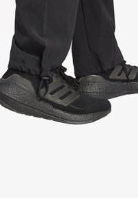 Schwarze Sportschuhe mit strukturierten Sohlen, ausgestattet mit Mesh-Obermaterial, drei Streifen-Akzenten und Schnürdesign. Die Hose hat einen umgeschlagenen Saum.
