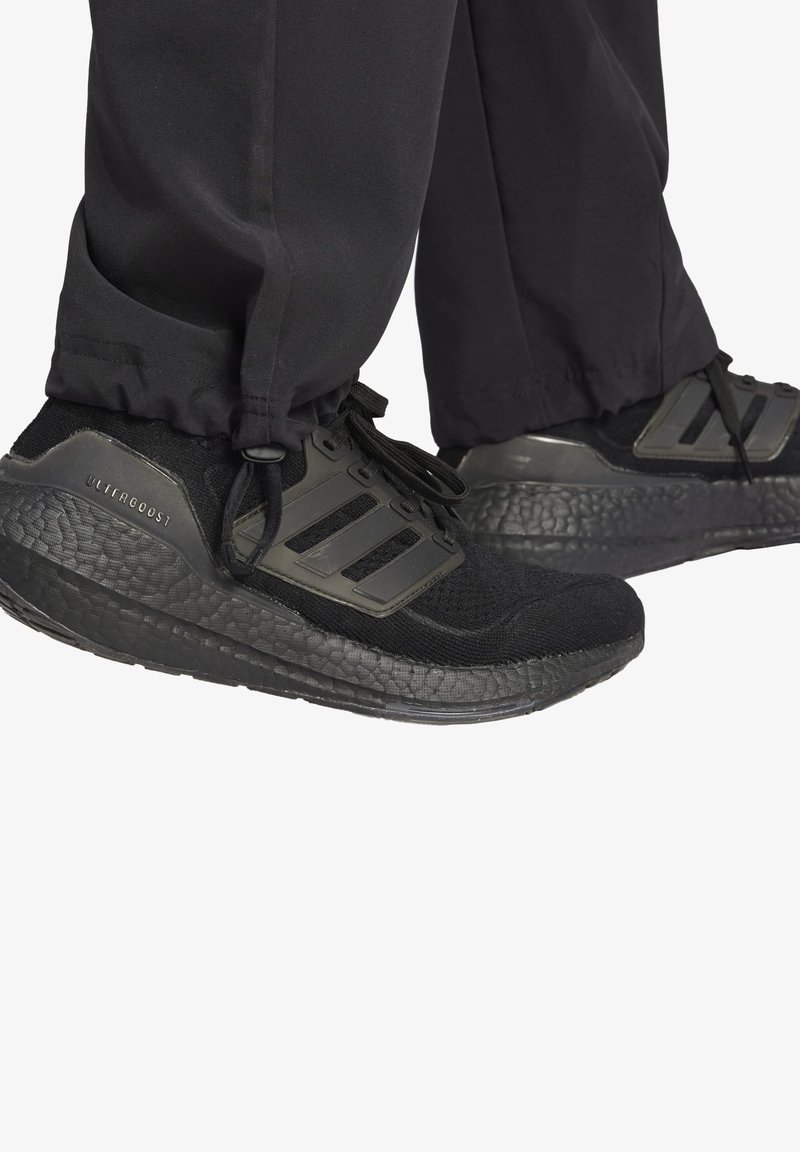 Zapatillas deportivas negras con suelas texturizadas, con parte superior de malla, acentos de tres franjas y diseño de cordones. Los pantalones tienen dobladillo acampanado.