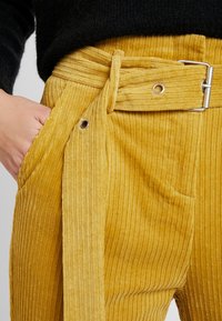 Gula corduroybyxor med bälte i midjan, sidofickor och vertikal ribbad textur. Silverdetaljer på bältet.