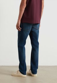 Jeans in denim di colore blu scuro, fit dritto; indossati con una camicia bordeaux e sneakers beige. I jeans presentano leggere abrasioni e tasche tradizionali.