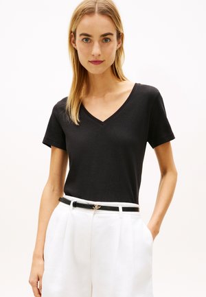 Donna che indossa una maglietta nera a scollo a V con maniche corte infilata in pantaloni bianchi a vita alta con cintura nera, in piedi con una mano in tasca.