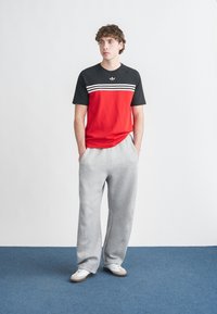 T-shirt de manga curta vermelha e preta com riscas brancas, combinada com calças de fato de treino cinza claro e ténis brancos, sobre um tapete azul.