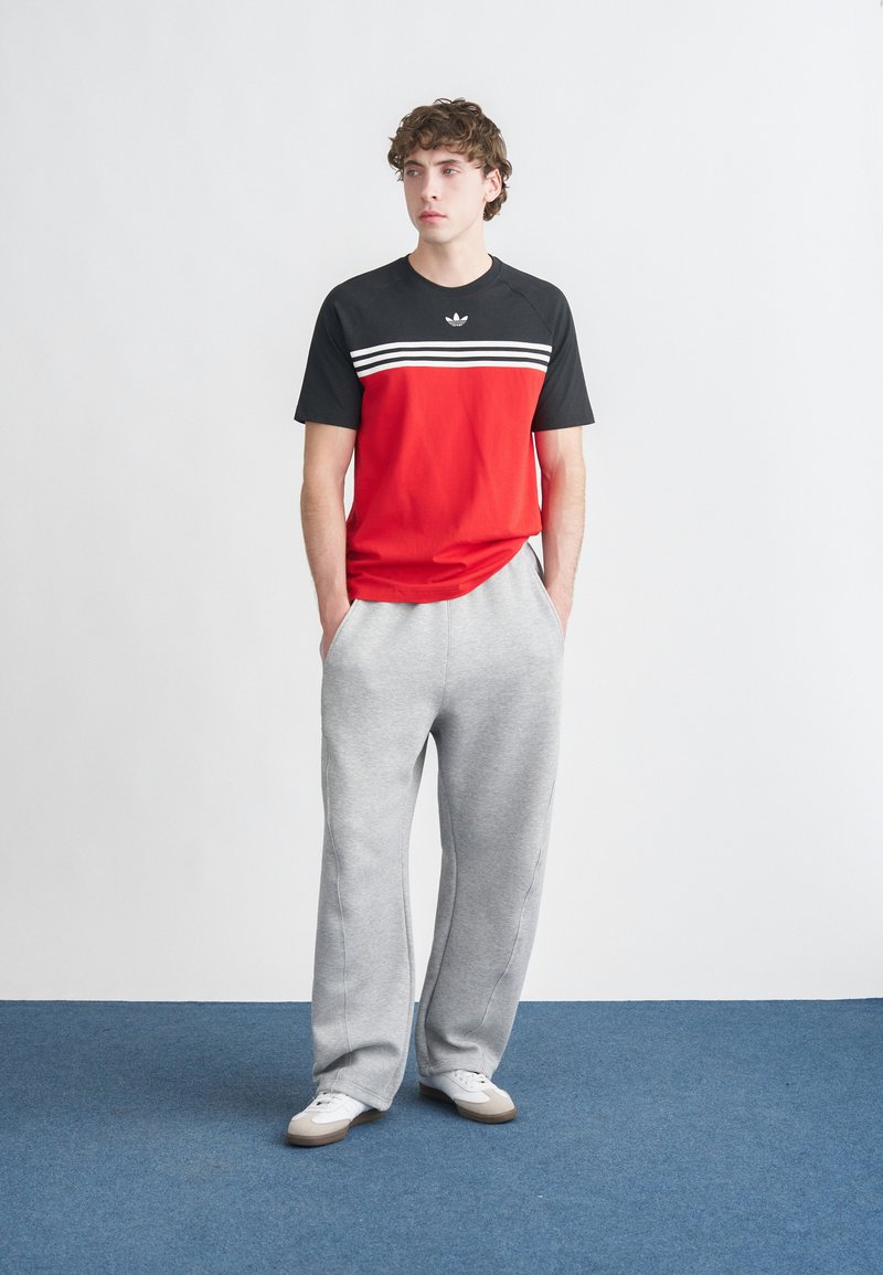 T-shirt à manches courtes rouge et noire avec des rayures blanches, associée à un pantalon de jogging gris clair et des baskets blanches sur un tapis bleu.