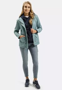 Jasnozielona wodoodporna kurtka z kapturem, z zamykanymi na zamek kieszeniami i regulowanym w pasie. Nosi ją na granatowym hoodie, w połączeniu z dopasowanymi dżinsami i sneakersami.