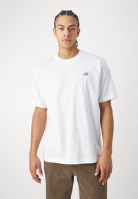 Jovem com cabelo trançado a usar uma t-shirt branca simples da New Balance e calças castanhas, de pé contra um fundo branco simples.