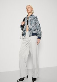 Veste bomber florale en tissu bleu et blanc avec des poignets côtelés. Associée à un pantalon large gris clair et des escarpins noirs à bout pointu.