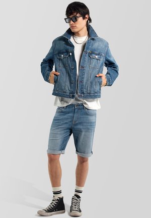 Giovane uomo con occhiali da sole, giacca di jeans a strati sopra camicia bianca, pantaloncini in jeans, calzini a righe e sneakers alte nere, in piedi.