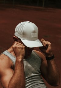 White cap with mesh detailing on the sides, flat brim, and a small logo patch on the front. Worn by a person in a gray tank top.
