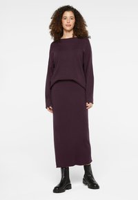 Lila gestricktes Set mit einem langärmligen Oberteil und einem passenden Maxirock. Sanfte Textur mit lockerer Passform. Kombiniert mit schwarzen Knöchelstiefeln.