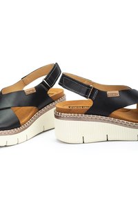 Sandalias de plataforma de cuero negro con diseño de tiras cruzadas, plantilla acolchada en tono marrón, suela blanca de contraste y detalles decorativos de costura.