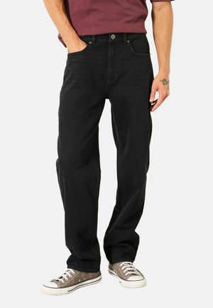 Mann trägt lockere schwarze Jeans, graue Leinenschuhe und ein bordeauxfarbenes Hemd, eine Hand in der Tasche vor weißem Hintergrund.