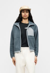 Γυναίκα που φοράει μπεζ καπέλο, μπλε φλις μπουφάν με φερμουάρ, μαύρο μπλουζάκι με λογότυπο και λευκό παντελόνι, στέκεται μπροστά σε ένα απλό φόντο.