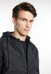 Sort zip-up jakke med hætte, der har en glat tekstur og en kontrastfarvet mørk inderside. Nærbillede af kraven og lynlåsdetaljen.