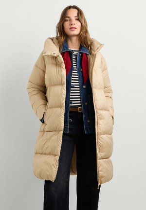 Frau steht und trägt einen beigen langen Steppmantel über einer blauen Jeansjacke und schwarzen Hosen, die Hände in den Manteltaschen, vor einem einfarbigen Hintergrund.