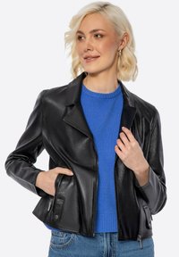 Schwarze Lederjacke mit tailliertem Design, Reverskragen, Reißverschluss vorne und Seitentaschen, getragen über einem blauen Pullover.