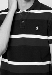 Proužkovaná polo košile v černobílé barvě s límečkem, třemi knoflíky a malým logem na levé straně hrudi.