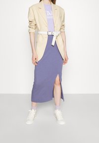 Béžový blazer přes levandulové šaty s bočním rozparkem, doplněný bílým páskem a světlými teniskami pro vrstvený, casual vzhled.