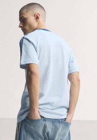 Jeune homme aux cheveux courts portant un t-shirt bleu clair avec des rayures bleu foncé sur les manches et un jean bleu, les mains dans les poches arrière.