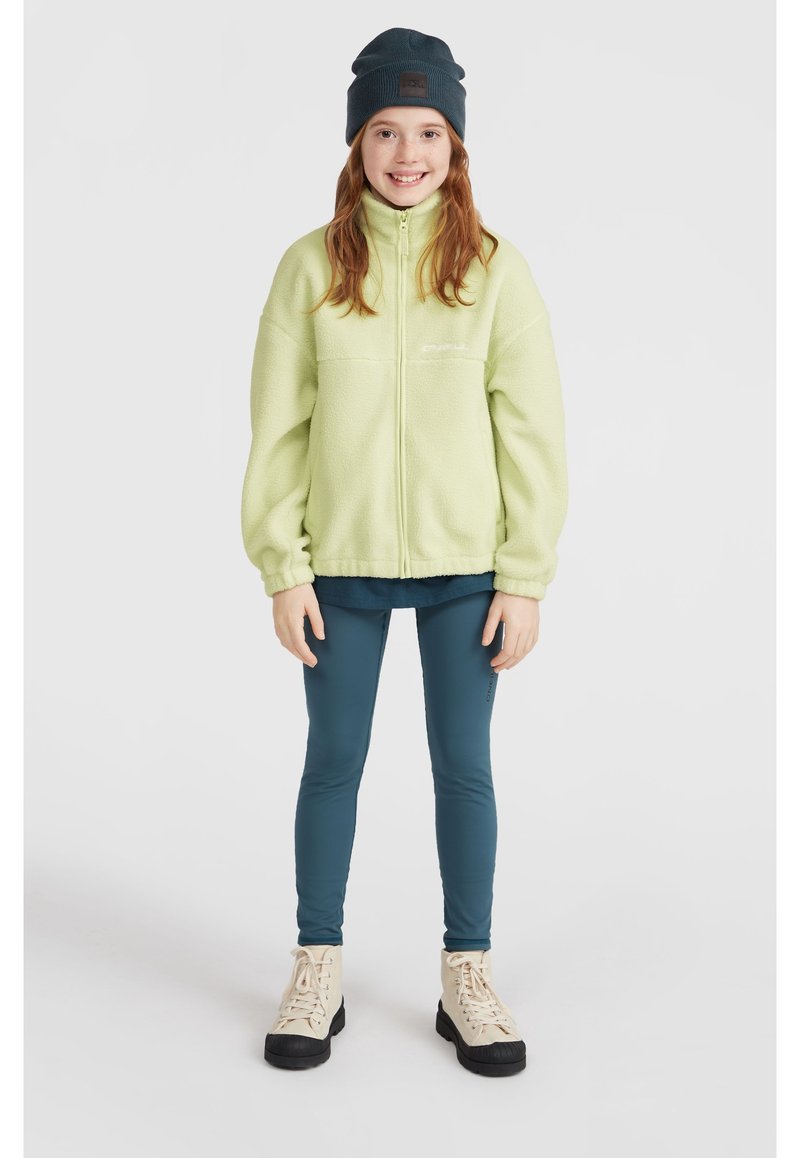 Grön fleecejacka med hög krage, marinblå mössa, teal leggings och ljusa ankelstövlar. Släta texturer, avslappnad passform, minimal varumärkeslogotyp.