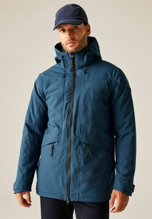 Mężczyzna noszący niebieską zimową kurtkę z kapturem, zamkiem błyskawicznym i kieszeniami z patkami, w ciemnoniebieskiej czapce i ciemnych spodniach, zwrócony przodem.