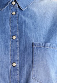 Ljusblå denimskjorta med spetsig krage, framknäppning och bröstficka. Har synliga sömmar och en slät textur.