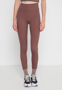 Magas derekú barna leggings rugalmas anyagból, sima tapintással és lapos varrással. Fekete és pink sportcipővel viselve.