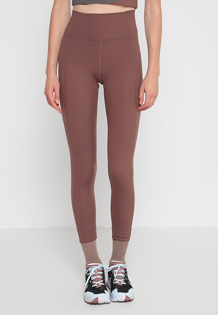 Magas derekú barna leggings rugalmas anyagból, sima tapintással és lapos varrással. Fekete és pink sportcipővel viselve.