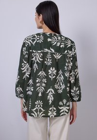 Blusa verde a fiori con disegni bianchi di piante, tessuto leggero, scollatura rotonda e maniche arricciate. Abbinata a pantaloni color crema.