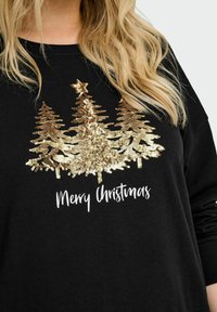 Crni dukserica s tri zlatne šljokaste božićne jelke i bijelim natpisom "Sretan Božić" koju nosi osoba s dugom plavom kosom.