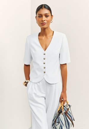 Femme en haut blanc à col en V boutonné et pantalon blanc ample, tenant un sac à main rayé avec des poignées en bambou, debout devant un fond uni.