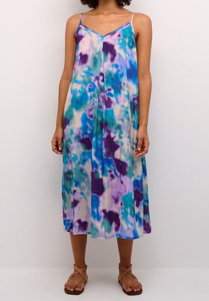 Femme portant une robe midi sans manches, ample, à motif tie-dye bleu, violet et bleu sarcelle, associée à des sandales à lanières marron clair.