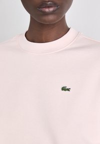 Világos rózsaszín pulóver puha anyagból, kerek nyakkivágással és kis zöld krokodil logóval a mellrészen, sima tapintással.