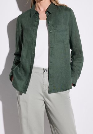 Vrouw draagt een groene blouse met lange mouwen en knopen over een witte top met lichtbeige broek, handen in de zakken tegen een effen achtergrond.