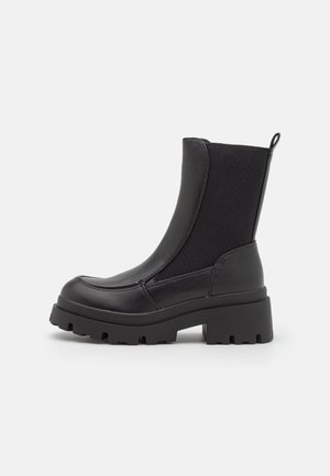 Bottes noires à la cheville avec une semelle en caoutchouc épaisse, combinant cuir lisse et tissu extensible. Languette d'attache à l'arrière ; design élégant et moderne.