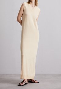 Robe en tricot sans manches de couleur beige avec un décolleté en V, et finitions côtelées au niveau du cou et de l'ourlet. Portée avec des sandales noires à lanières, debout sur un sol neutre.