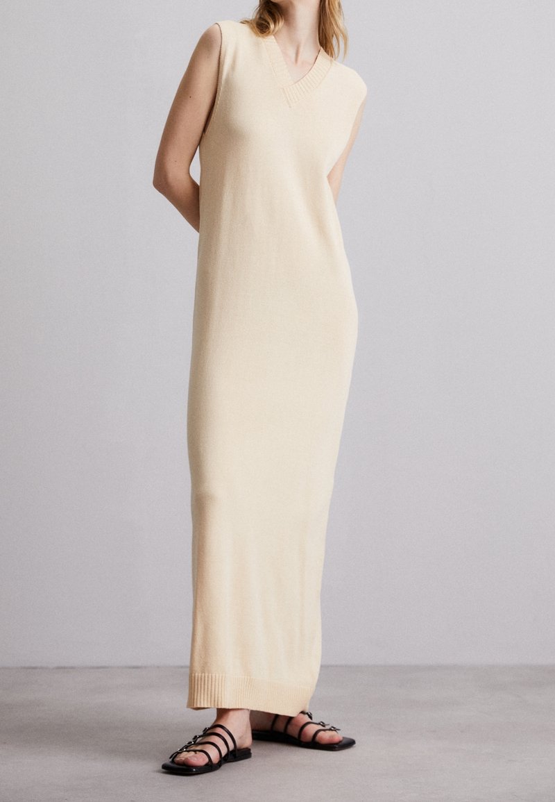 Robe en tricot sans manches de couleur beige avec un décolleté en V, et finitions côtelées au niveau du cou et de l'ourlet. Portée avec des sandales noires à lanières, debout sur un sol neutre.