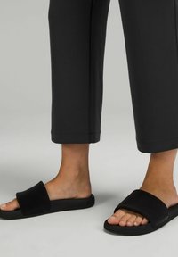 Fötter i svarta slidesandaler och svarta cropped byxor, stående på en neutral ljusgrå yta.