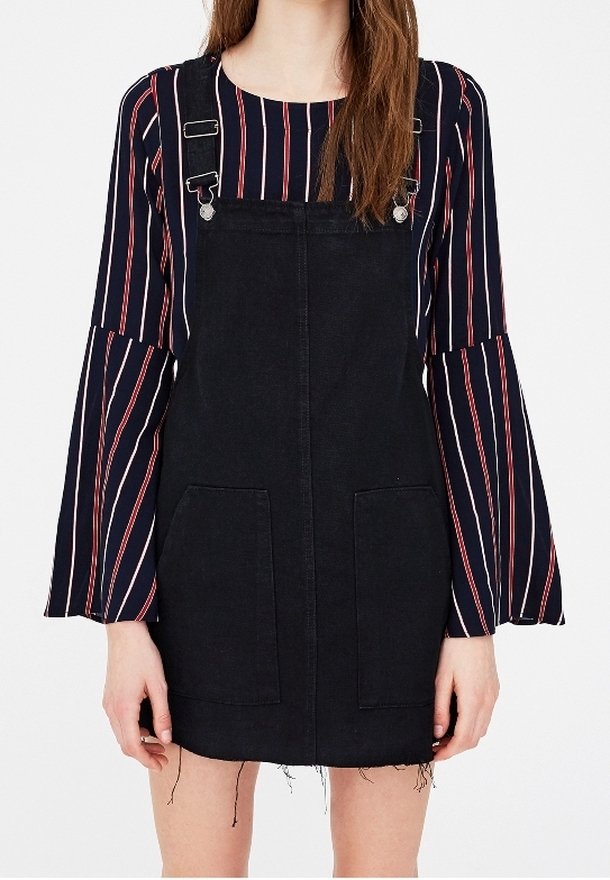 Femme portant une robe salopette en denim noir sur un chemisier bleu marine à manches longues avec des rayures verticales rouges et blanches.