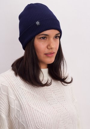 Navy ribbed knit beanie with a logo patch, worn by a person with dark hair. Textured white sweater partially visible.