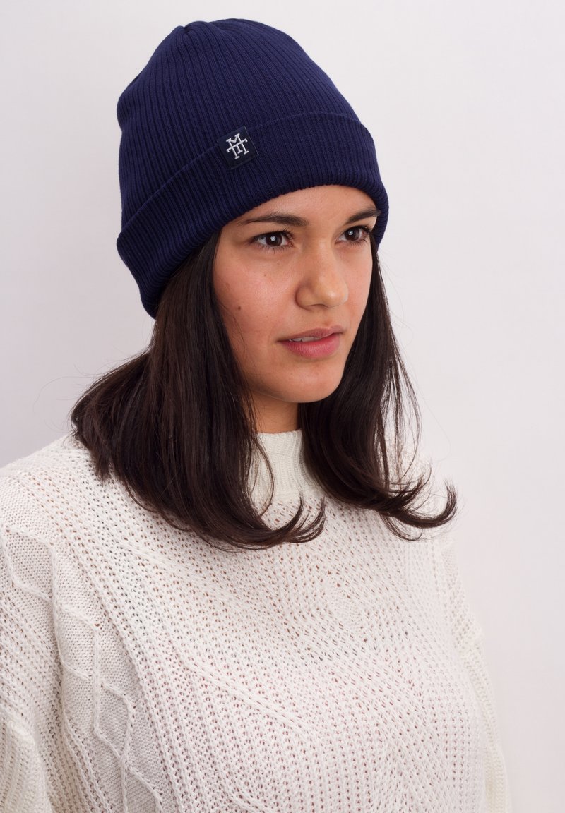 Navy ribbed knit beanie with a logo patch, worn by a person with dark hair. Textured white sweater partially visible.