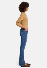 Beige gebreid sweater, blauwe flared jeans en flats met luipaardprint. Zijaanzicht benadrukt de getailleerde silhouet en textuur van de materialen.