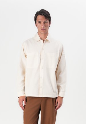 Camisa de botón de color beige claro con cuello, dos bolsillos en el pecho, mangas largas y dobladillo recto. Llevada sobre pantalones marrones.