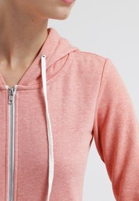 Mujer vestida con una sudadera con cremallera de color rosa claro y cordones blancos, mostrada desde los hombros hasta la barbilla sobre un fondo liso.