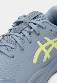 Svetlo modra športna čevlja z mrežasto teksturo, ki ima teksturirano površino, rumene logotipne akcentne in sivo vezalko.