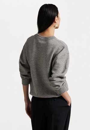 Kvinde med langt sort hår iført en grå sweater og sorte bukser, stående med den ene hånd i lommen, vendt væk mod en hvid baggrund.