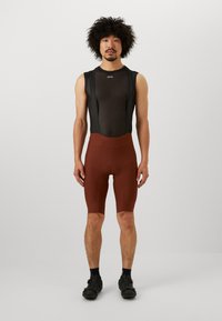 Herren-Radsportoutfit mit einem schwarzen Mesh-Oberteil im ärmellosen Design und braunen gepolsterten Shorts, gefertigt aus dehnbarem, feuchtigkeitsableitendem Material.